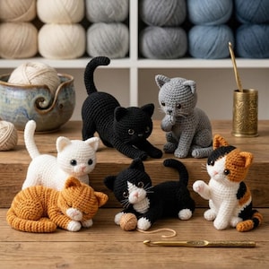 Op de afbeelding: Een verzameling van zes gehaakte kattenfiguurtjes in diverse kleuren, waaronder zwart, wit, grijs, oranje en lapjeskat. De katten staan in verschillende houdingen, zoals zittend, uitrekkend of liggend, vergezeld door een klein bolletje wol.