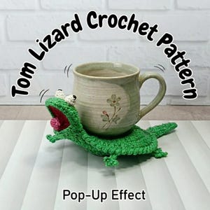 May include: A green crocheted lizard with a wide-open mouth and white eyes, serving as a base for a light green ceramic mug with a floral design. The text reads Tom Lizard Crochet Pattern and Pop-Up Effect in black lettering.