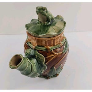 May include: A ceramic teapot featuring a brown bamboo-textured body and a green lid topped with a sculpted frog sitting on a lily pad. The spout is shaped like a green leaf, and the overall design incorporates natural elements in shades of brown and green.