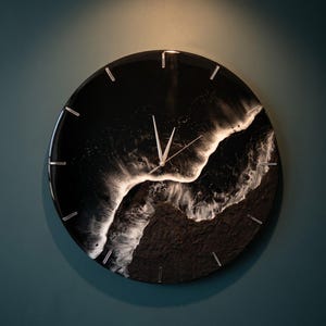 May include: A circular wall clock featuring a glossy black surface with white resin accents that mimic crashing waves. The clock face includes thin silver hour markers and slender silver hands, with a textured black section at the bottom.