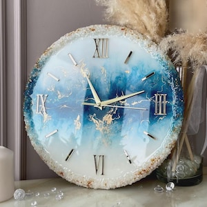 May include: A round wall clock featuring a swirling blue and white resin face with gold leaf accents. The clock has silver Roman numerals at the twelve, three, six, and nine o'clock positions, with silver hour markers and thin silver clock hands.