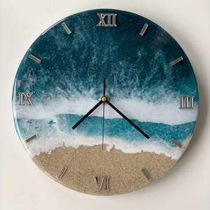May include: A round wall clock featuring a resin art design that mimics an ocean shoreline. The clock face displays a sandy beach at the bottom, transitioning into turquoise water with white sea foam waves, and deep blue water above. It has silver Roman numerals.