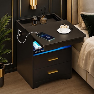 May include: A black bedside table with two drawers featuring gold handles, a top surface with a built-in charging port, a smartphone connected to a white charging cable, a white coffee cup on a saucer, and an open shelf with blue LED lighting.