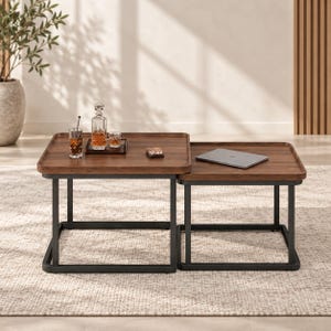 May include: A set of two nesting coffee tables featuring dark brown wooden square tops with raised edges and black metal frames. The larger table holds a glass decanter and two glasses, while the smaller table supports a closed silver laptop.
