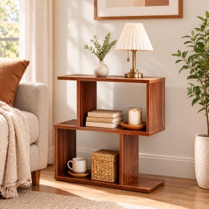 May include: A dark wood S-shaped bookshelf with two levels. The top shelf holds a white vase with green foliage, a brass lamp with a pleated shade, a stack of three books, and a candle in a glass jar. The bottom shelf holds a mug and a woven box.
