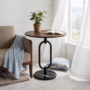 May include: A round side table with a dark wood top, a black metal oval-shaped pedestal base, and a circular black marble base. The table features an open center design in the pedestal and is positioned next to a chair with a light blue throw.