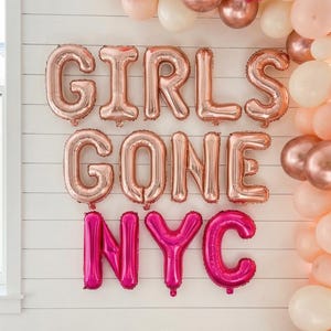 May include: A festive scene with rose gold and pink balloons forming an arch and the words "GIRLS GONE NYC". A table holds champagne, glasses, books, and a candle, creating a celebratory atmosphere for a party or event.