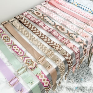 May include: A collection of colorful woven straps with gold metal hardware. The straps are arranged in a row on a white surface. The straps are all different colors and patterns, including pink, red, brown, beige, and green.