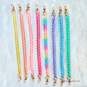 May include: A collection of colorful acrylic chain straps with gold-tone clasps. The straps are in various shades, including yellow, orange, pink, red, rainbow, purple, blue, and green. The chains are displayed on a white, fluffy surface.
