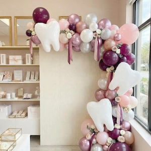 Puede incluir: Un arco de globos con temática dental, con globos blancos en forma de diente y una mezcla de globos rosas, burdeos y blancos. El arreglo incluye acentos florales y cintas, creando una exhibición festiva.