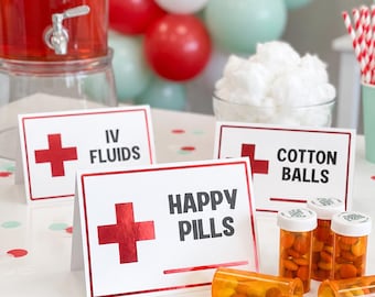 May include: Three white rectangular signs with red borders and red cross symbols. The signs are labeled IV FLUIDS, COTTON BALLS, and HAPPY PILLS in black text. Several orange plastic pill bottles filled with colorful candies sit nearby.