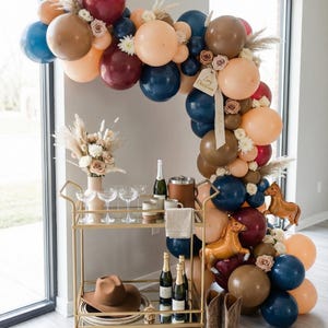 May include: A festive party scene with a gold bar cart, balloons, and decorations. The balloon arch features shades of blue, brown, and peach. A brown cowboy hat and boots are on display, along with champagne and glasses.