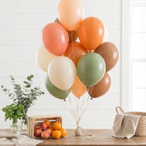 May include: A bouquet of balloons in various colors, including peach, orange, cream, green, and brown. The balloons are tied together with twine. A wooden crate filled with peaches and oranges sits on a wooden table, along with a basket and greenery.