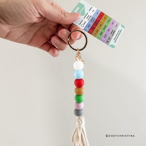 May include: A colorful beaded keychain with a white tassel. The beads are white, light blue, red, brown, green, light purple, and gray. The keychain is attached to a gold ring.
