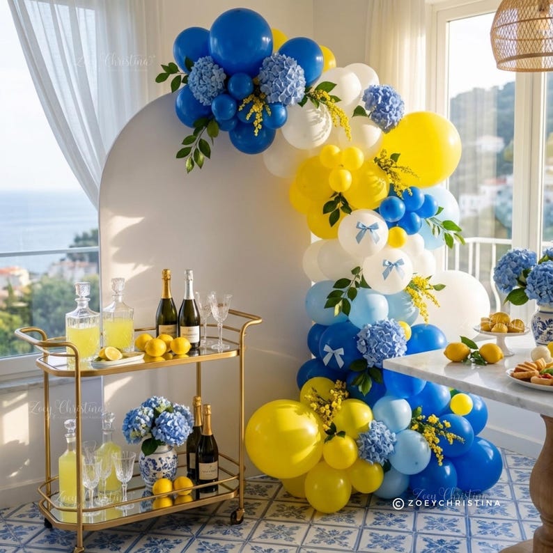 May include: A festive party scene with a gold bar cart, champagne, and lemons. A balloon arch in blue, yellow, and white with floral accents decorates the space. The floor has a blue and white tile pattern.