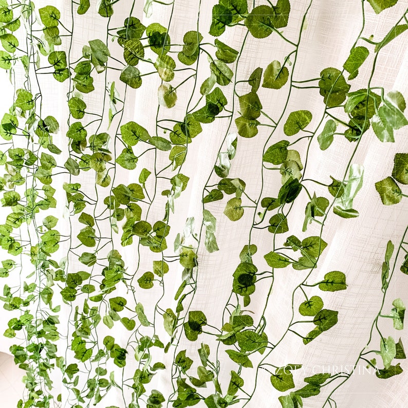 May include: A white sheer curtain with a green artificial vine garland hanging in front of it. The vine garland has many small green leaves.