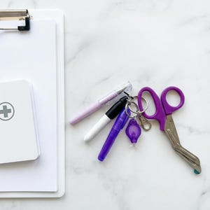 May include: A medical-themed flat lay featuring a white clipboard, a small first aid kit, and several pens. A set of purple medical shears and a small keychain light are also present. The items are arranged on a white marble surface.