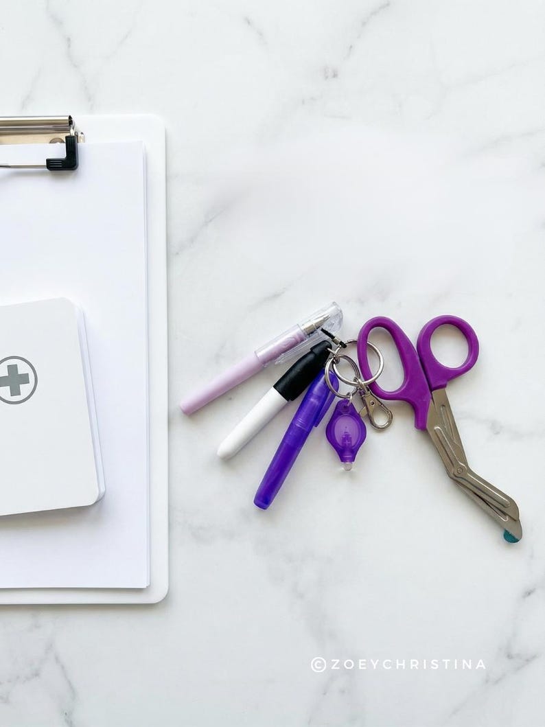 May include: A medical-themed flat lay featuring a white clipboard, a small first aid kit, and several pens. A set of purple medical shears and a small keychain light are also present. The items are arranged on a white marble surface.