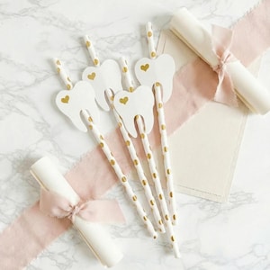 May include: White paper straws with gold polka dots and tooth-shaped toppers with gold heart accents. Rolled paper scrolls tied with pink ribbon are also present. The items are arranged on a marble surface.