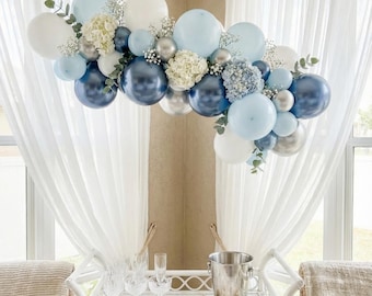 May include: A decorative balloon arch in shades of blue, white, and silver, adorned with white flowers and greenery. The arrangement is suspended above a white serving cart with champagne flutes and a bucket.