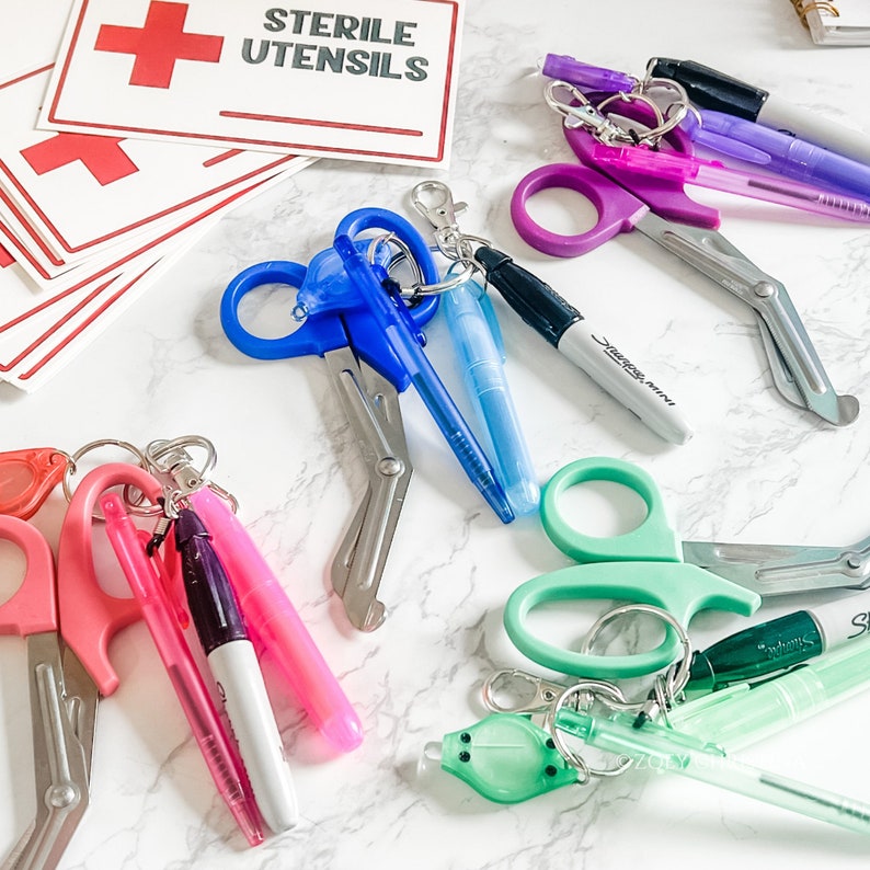 May include: Assorted medical shears in pink, blue, purple, and green, attached to keychains with pens and a mini Sharpie. The image also includes red cross symbols and a sign that reads "STERILE UTENSILS."