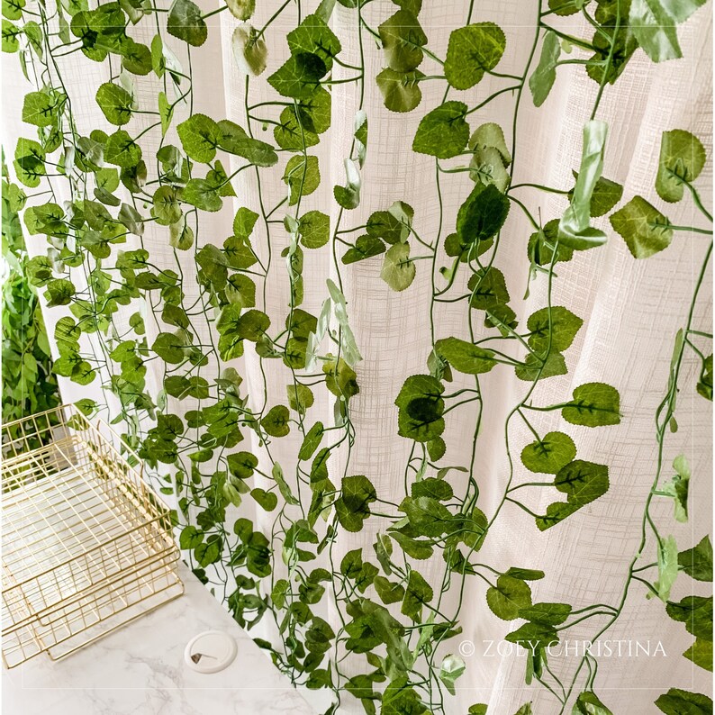 May include: A white sheer curtain with a green artificial vine garland draped over it. The garland features realistic-looking leaves and stems.