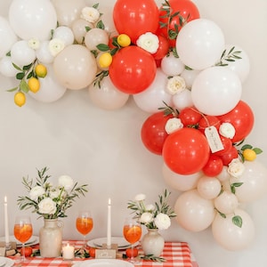 May include: A festive scene with a red and white checkered tablecloth, set with plates, glasses, and candles. A balloon arch of red, white, and cream balloons is decorated with lemons and flowers. A tag reads "Merry Christmas."
