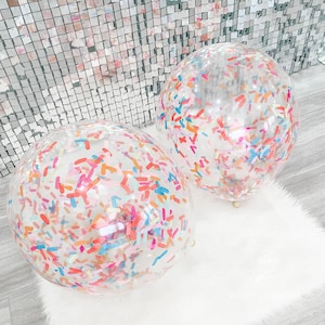 May include: Two clear balloons filled with colorful confetti. The confetti is in the shape of sprinkles and is in shades of pink, orange, blue, and yellow.