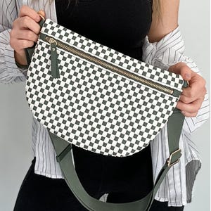 May include: A checkered pattern fanny pack with a gold zipper and olive green strap. The bag features a black and white checkerboard design and is held by a person. The bag has a gold buckle and a zipper pull.