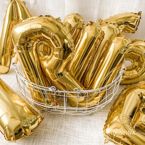 May include: A group of gold foil balloons in a wire basket. The balloons are shaped like letters and are arranged in a random order.