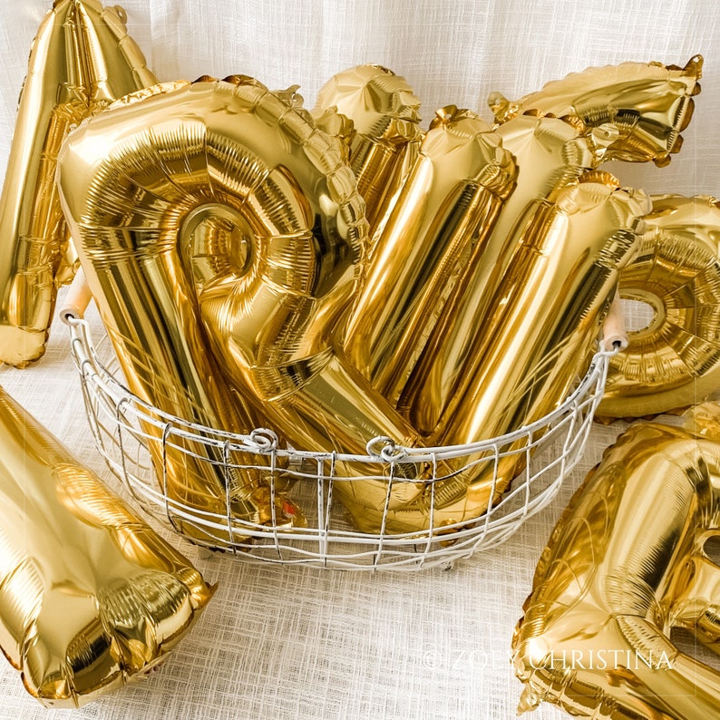 May include: A group of gold foil balloons in a wire basket. The balloons are shaped like letters and are arranged in a random order.