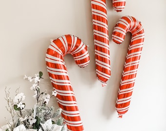 May include: Three red and white striped candy cane shaped balloons. The balloons are hanging on a white wall.