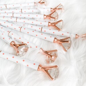 May include: A group of white pens with pink polka dots and rose gold diamond-shaped tops. The pens are lying on a white fluffy surface.