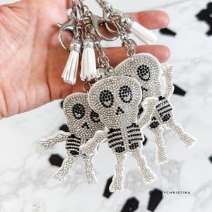 May include: Silver skeleton keychains with white tassels. Each skeleton is covered in sparkling rhinestones with black accents. The keychains are attached to silver metal clasps and chains. The keychains are a Halloween or gothic theme.