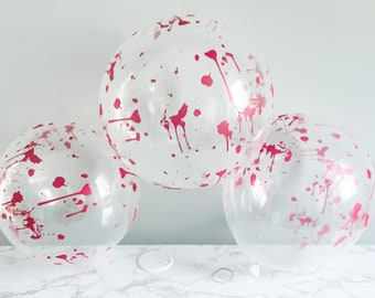 May include: Three clear balloons with a pink splatter design. The balloons are round and inflated, with the pink paint splatters adding a decorative touch. The balloons are on a white marble surface.