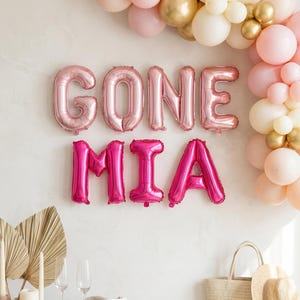 May include: Pink foil balloons spelling "GONE MIA" are displayed on a white wall, with a balloon arch in the background. A table is set with champagne flutes, candles, and a straw bag with a hat.