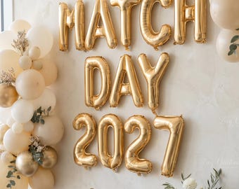 May include: Gold foil balloons spelling MATCH DAY 2027 arranged on a wall with clusters of cream and metallic gold balloons, a bottle of champagne, four clear glasses, and a sign reading Do I Now? Matched! next to a vase of white flowers.
