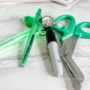 May include: A collection of green stationery items, including a pen, highlighter, scissors, and a Sharpie marker. The items are arranged on a white and gray marble surface. The Sharpie marker is white with a black cap.