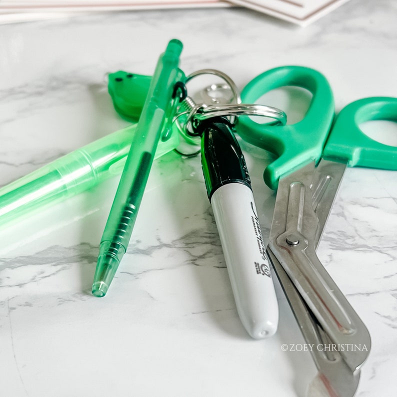 May include: A collection of green stationery items, including a pen, highlighter, scissors, and a Sharpie marker. The items are arranged on a white and gray marble surface. The Sharpie marker is white with a black cap.