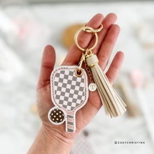 May include: A white and gray checkered paddleball keychain with a pink and brown ball attached. The keychain has a gold key ring and a cream-colored tassel with a gold charm that says "T".