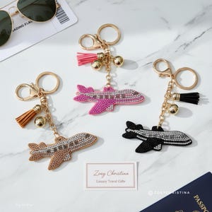 May include: Three airplane keychains with gold-colored hardware, each featuring a different color airplane: brown, pink, and black. Each keychain has a tassel and a small gold bell. The keychains are embellished with rhinestones.