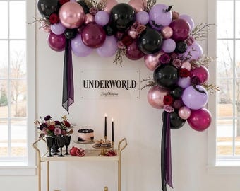 May include: A balloon arch in shades of black, metallic rose gold, deep burgundy, and lavender, accented with dried branches and small black bat cutouts. A sign on the wall reads UNDERWORLD. A gold bar cart holds a cake, candles, and glassware.
