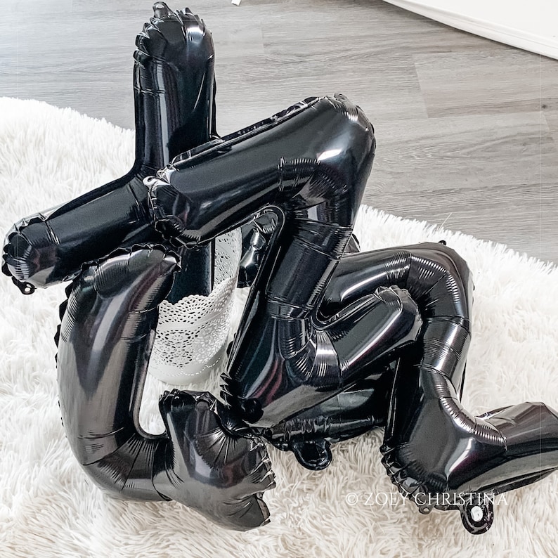 May include: Black balloon letters spelling out the word "LOVE" in a modern, abstract style. The balloons are arranged in a cluster, creating a dynamic and eye-catching display.