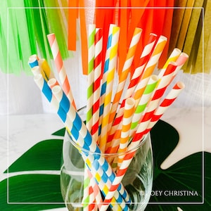 May include: A glass vase filled with colorful striped paper straws. The straws are red, yellow, blue, green, and pink. The vase is sitting on a green leaf.