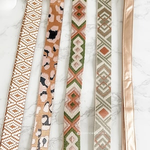 May include: A collection of patterned bag straps in various designs and colors. The straps include geometric, animal print, and solid satin styles. Colors include brown, tan, green, and peach. The straps are displayed on a white marble surface.