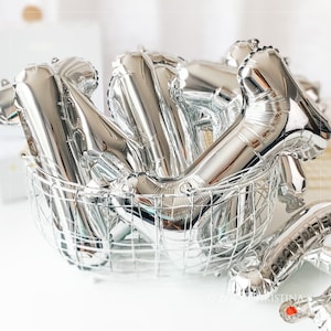 May include: A silver wire basket filled with silver foil letter balloons. The balloons are in the shape of the letters "N" and "Y".