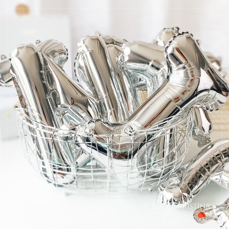 May include: A silver wire basket filled with silver foil letter balloons. The balloons are in the shape of the letters "N" and "Y".
