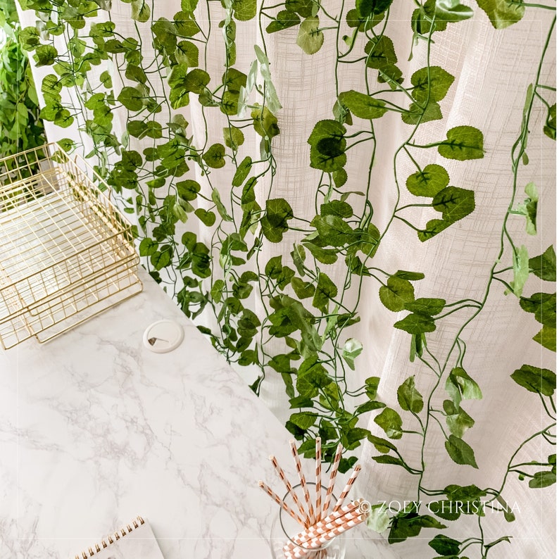 May include: A white sheer curtain with a green leafy garland hanging in front of it. The curtain is textured and has a subtle pattern. The garland is made of artificial leaves and vines.