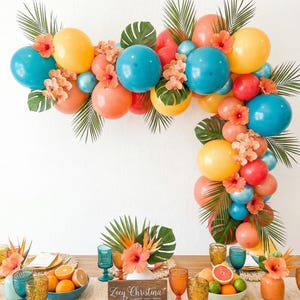 May include: A tropical-themed party table setting with a balloon arch in shades of blue, yellow, and orange, accented with tropical flowers and greenery. The table is set with colorful glassware, fruit, and place settings.