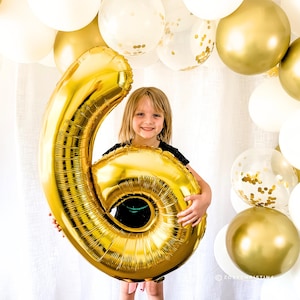 May include: A young girl holds a large gold number six balloon in front of a white background. The balloon is decorated with gold confetti. The background is decorated with white and gold balloons.